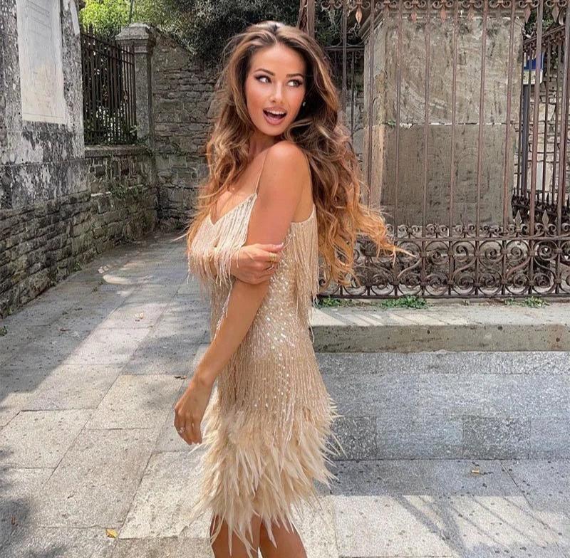 Woman in a beige fringe dress standing outdoors with a stone wall and metal gate in the background.